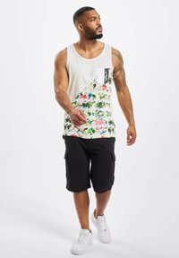 Herretanktop med hvid, spraglet stof og farverigt blomstermønster. Har en venstre brystlomme. Parret med sorte cargo-shorts og hvide sneakers.