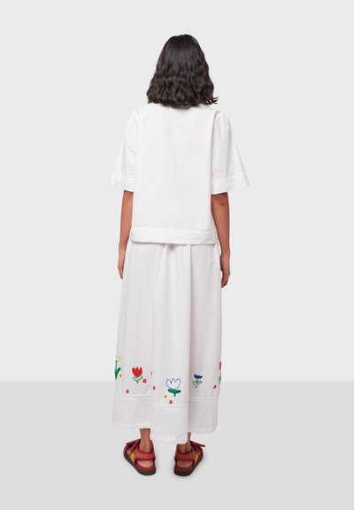 Witte katoenen outfit met korte mouwen en een lange rok. De rok heeft kleurrijke bloemenborduring aan de zoom en wordt gedragen met rode sandalen.