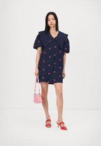 Τζιν φόρεμα με μεγάλο, φουσκωτό γιακά, κοντά φουσκωτά μανίκια και ροζ σχέδια καρδιάς. Στυλιζαρισμένο με κόκκινα τακούνια και μια μικρή ροζ τσάντα.