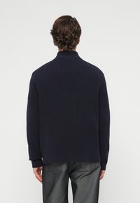 Pull en maille bleu marine avec un col haut, présentant une texture côtelée et une coupe décontractée. La vue de dos montre un minimalisme des détails et des manches longues.