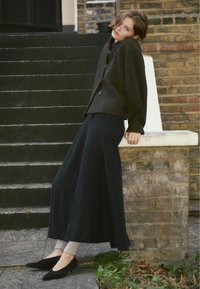 Tmavý kabát z vlněné směsi má velké knoflíky a je kombinován s dlouhou tmavou sukní. Viditelné jsou černé lodičky na špičce a šedé ponožky.