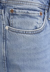 Los jeans de mezclilla azul claro presentan un bolsillo frontal con remaches de cobre, costuras en contraste y una superficie texturizada. La tela muestra un ligero desgaste.