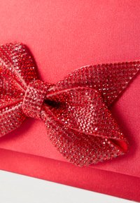 Crvena torbica s velikom teksturiranom mašnom ukrašenom blistavim crvenim šljokicama na glatkoj satenskoj podlozi.