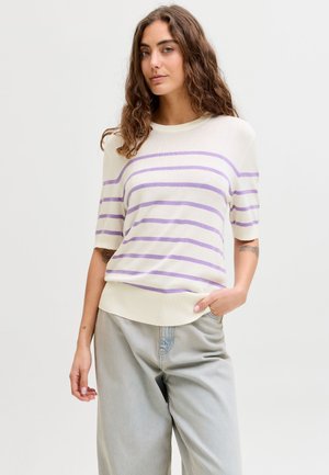 Jeune femme aux cheveux bruns bouclés portant un haut blanc à manches courtes avec des rayures violettes et un pantalon ample gris clair sur un fond uni.