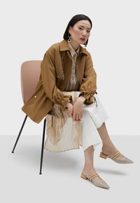 Brun jacka med puffärmar, beige randig skjorta, vita vida byxor och beigefärgade spikade mules. Sitter på en svart stol.