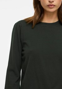 Top tricot verde închis cu mânecă lungă, guler rotund și detalii ușor bufante la umeri, având o textură fină și moale.