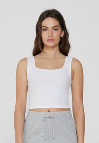 Weißes, geripptes Cropped Top mit quadratischem Ausschnitt und breiten Trägern, kombiniert mit grauen Sweatpants mit Kordelzug. Glatte Textur, einfaches Design.