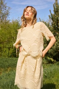 Femme debout en plein air, les yeux fermés, vêtue d'une robe au crochet crème avec des motifs circulaires, les mains dans les poches, sous un ciel bleu clair.