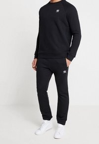 Man i svart Adidas-tröja och matchande joggingbyxor med vita Adidas-loggor, kombinerat med vita sneakers, står framför en enfärgad bakgrund.
