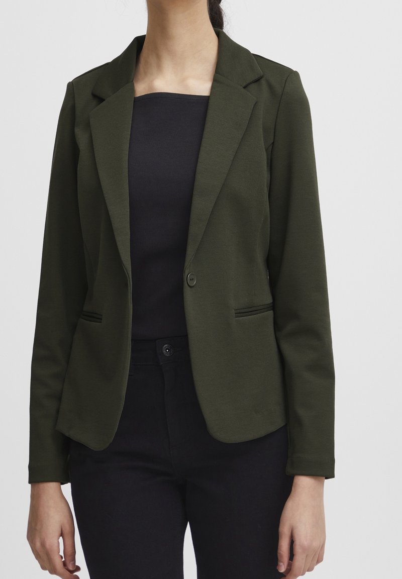 Femme portant un blazer vert foncé ajusté sur un haut noir et un pantalon noir, sur un fond clair uni.