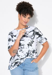 Kortærmet hvid bluse med sort blomstermønster, blødt stof og en subtil buet kant. Bæres med lyseblå jeans prydet med perler.