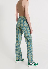 Débardeur marron ajusté avec un pantalon large à motifs bleus et jaunes présentant un design géométrique et des poches plates à l'avant, associé à des baskets vertes.