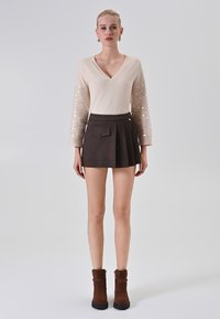 Donna che indossa un maglione beige a scollo a V con maniche di paillettes, una mini gonna plissettata marrone scuro con tasca e stivali alla caviglia marroni, in piedi su uno sfondo neutro.