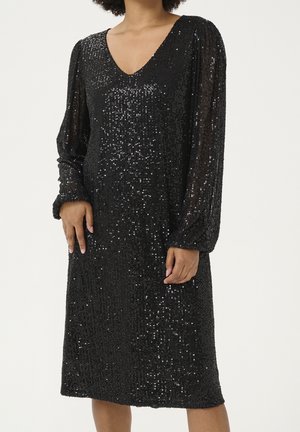 Femme portant une robe noire à sequins mi-longue avec des manches longues en voile transparent bouffantes et un décolleté en V, sur un fond uni.
