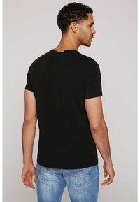 Camiseta de algodón negra con mangas cortas y cuello redondo, vista desde la parte posterior. El diseño tiene una pequeña etiqueta de acento en el cuello.