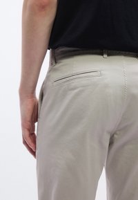 Béžové bavlnené nohavice s hladkým povrchom, vybavené jedným zadným vreckom a pútkami na opasok, zobrazené z boku s rukou odpočívajúcou v blízkosti.