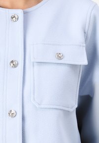 Ανοικτό μπλε μπουφάν από μαλακό ύφασμα με μεταλλικά κουμπώματα και δύο τσέπες μπροστά στο στήθος. Υφή επιφάνειας και δομημένος σχεδιασμός.