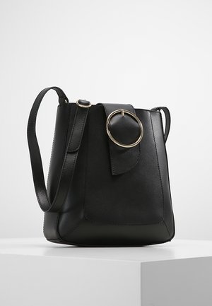 Bolso tipo cubo de cuero negro con una correa de hombro ajustable y una hebilla circular dorada en la solapa frontal, exhibido sobre un pedestal blanco.