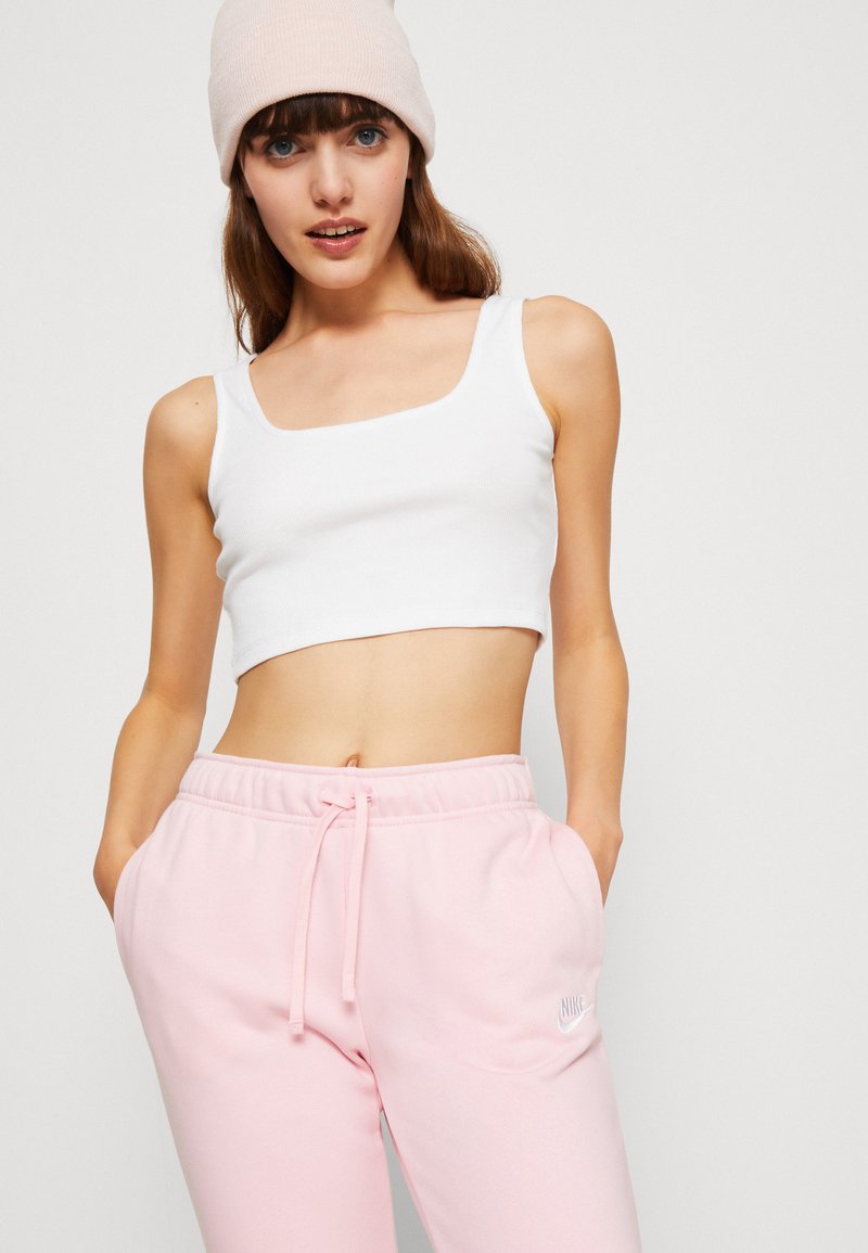 Jeune femme portant un haut court blanc, un pantalon de jogging rose clair de Nike et un bonnet rose clair, se tenant avec les mains dans les poches.