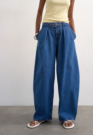 Persona que lleva vaqueros azules de tiro alto y pierna ancha con cinturón, una camiseta sin mangas de color amarillo claro, pulseras plateadas y sandalias blancas con tiras.