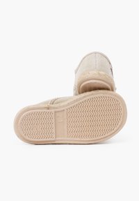 Chaussure slip-on beige tissée avec semelle texturée, présentant un bout arrondi et un subtle détail de logo près de l'ouverture. Taille marquée comme 21.