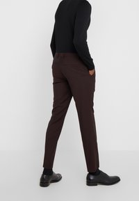 Uomo che indossa pantaloni scuri fitted marroni, maglia nera a maniche lunghe, calzini neri e scarpe eleganti nere, in piedi con le mani in tasca di fronte a uno sfondo bianco.