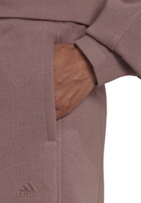 Jasnofioletowy sweter z ribbowym mankietem, z boczną kieszenią i subtelnie wytłoczonym logo Adidas w pobliżu dołu. Miękka tekstura.