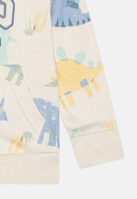 Sweatshirt beige clair avec des motifs de dinosaures colorés en bleu, jaune et vert, doté d'une poche kangourou et de poignets côtelés.