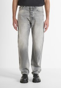 Hombre que lleva jeans rectos de color gris desgastado y botas negras de cuero con cordones, de pie frente a un fondo blanco liso.