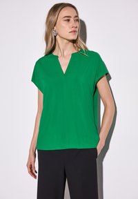 Top verde a maniche corte con scollatura a V. Realizzato in tessuto morbido, presenta una vestibilità rilassata e un orlo dritto. Abbinato a pantaloni neri.