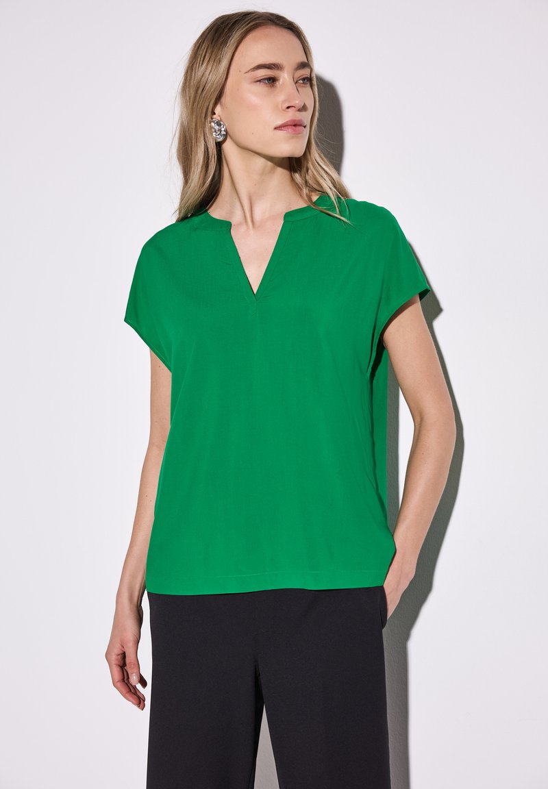 Top verde a maniche corte con scollatura a V. Realizzato in tessuto morbido, presenta una vestibilità rilassata e un orlo dritto. Abbinato a pantaloni neri.