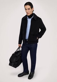 Svart jacka med krage, blå och vit randig skjorta, mörka jeans och svarta skor. Håller en svart väska. Mjuk tygtextur.