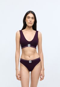 Braletă violet cu nervuri, cu bretele late și decolteu adânc în formă de V, având un patch logo și bandă rayată. Slip violet asortat cu un design similar.