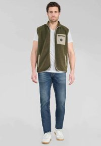Gilet in pile verde con tasca frontale color crema, apertura frontale e due tasche laterali, abbinato a jeans blu e sneakers bianche.