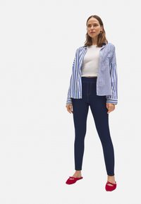 Camisa abotonada a rayas azul y blanca superpuesta sobre un top corto blanco, combinada con leggings azul oscuro y zapatos rojos tipo slip-on.