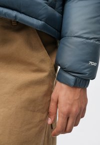 Pravá ruka osoby oblečenej v modrej nafúkanej bunde s nápisom "700" na manžete a hnedých štruktúrovaných nohaviciach na bielom pozadí.