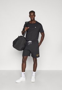 Μαύρο αθλητικό κοντομάνικο μπλουζάκι και σορτς, λευκό κορδόνι, κεντημένο λογότυπο, συνοδευόμενα από λευκά αθλητικά παπούτσια και μαύρη τσάντα duffel.