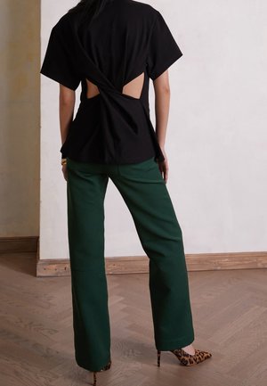 Femme debout à l'intérieur portant un haut noir à manches courtes avec un dos ouvert torsadé, un pantalon large vert et des talons hauts imprimés léopard.