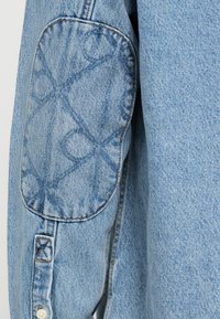 Camisa de denim azul claro com um remendo cosido no cotovelo com um padrão circular, punho abotoado com um detalhe em X e um acabamento texturizado.