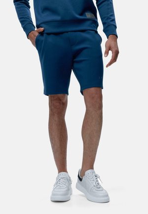 Pantaloni corti in felpa blu scuro con una forma sagomata, tasche laterali e dettagli di branding discreti. Abbinati a scarpe da ginnastica bianche con accenti blu navy.