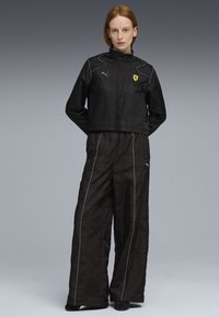 Jachetă neagră, scurtă, cu guler înalt și detalii gri, având logo-ul galben Ferrari. Asortată cu pantaloni negri, largi, cu umplutură, având accente albe.