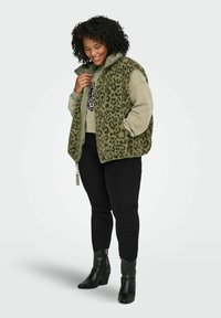 Smilende kvinne iført olivengrønn fleecevest med leopardmønster over matchende genser, svarte bukser og svarte ankelstøvletter med hæl.