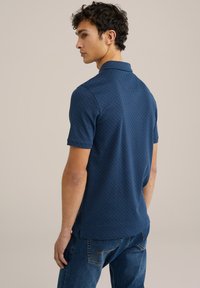 Tamsiai mėlyna polo marškinėliai su geometriniu raštu, trumpomis rankovėmis ir klasikiniu apykakle, derinami su mėlynais džinsais.