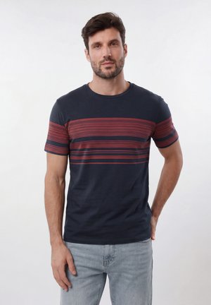 Rövid sötét hajú, szakállas férfi, aki sötétkék pólót visel piros csíkokkal és világoskék farmert, egyszínű fehér háttér előtt áll.