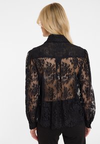 Schwarze Spitzbluse mit transparentem Rücken, floralen Mustern, langen Ärmeln und einem kragenähnlichen Halsauschnitt. Der Stoff ist zart und hat strukturierte Akzente.