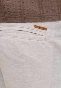 Gros plan de la poche d'un pantalon beige avec une petite étiquette marron de la marque Jack & Jones cousue près du bord supérieur sous un tissu marron texturé.