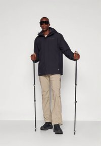 Mann som bærer svart jakke, beige turluer, svarte sko og solbriller, og holder svarte turstaver mot hvit bakgrunn.