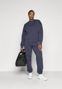 Marineblå hettegenser og matching joggebukser, laget av myk stoff. Svart duffelbag holdt i den ene hånden; modellen har på seg hvite sneakers.