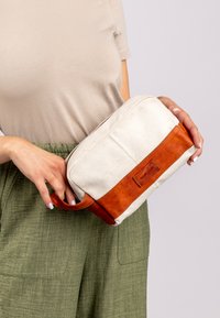 Kosmetyczka z płótna z brązową skórzaną podstawą i uchwytem. Prostokątny kształt, kremowy kolor z teksturowaną powierzchnią i naszywką z logo.