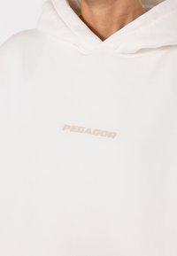 Sweat à capuche blanc en tissu doux, avec une coupe décontractée. Logo "PEGADOR" en ton beige clair, situé sur la poitrine.
